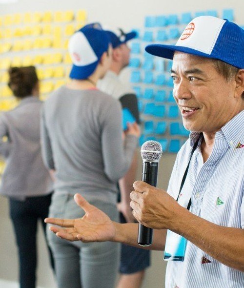 A facilitator with a microphone at an Offers and Needs Market, with participants placing sticky notes on a wall